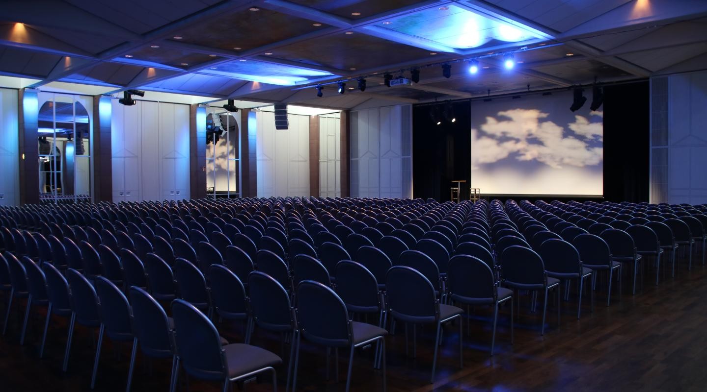 Schlossgartenhalle Reihenbestuhlung Schlossgartenhalle mit Reihenbestuhlung, Leinwand in der Mitte auf der Bühne und einem Stehtisch neben der Leinwand