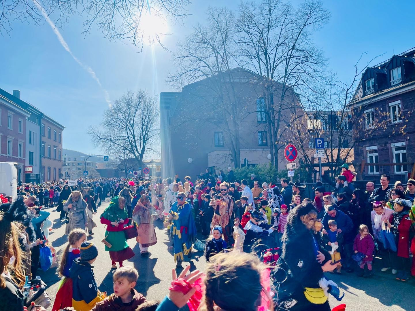 Rosenmontagsumzug Ettlingen Eine festliche Straßenszene mit vielen Menschen in Kostümen, die sich für einen Umzug oder eine Parade versammelt haben. Die Sonne scheint hell am Himmel, und viele Zuschauer stehen am Straßenrand, einige mit Kindern. Gebäude und Bäume umgeben die Straße.