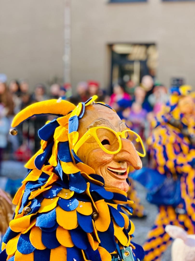 Narr in blau-gelbem Fleckenkostüme mit Holzmaske und einer gelben Brille auf
